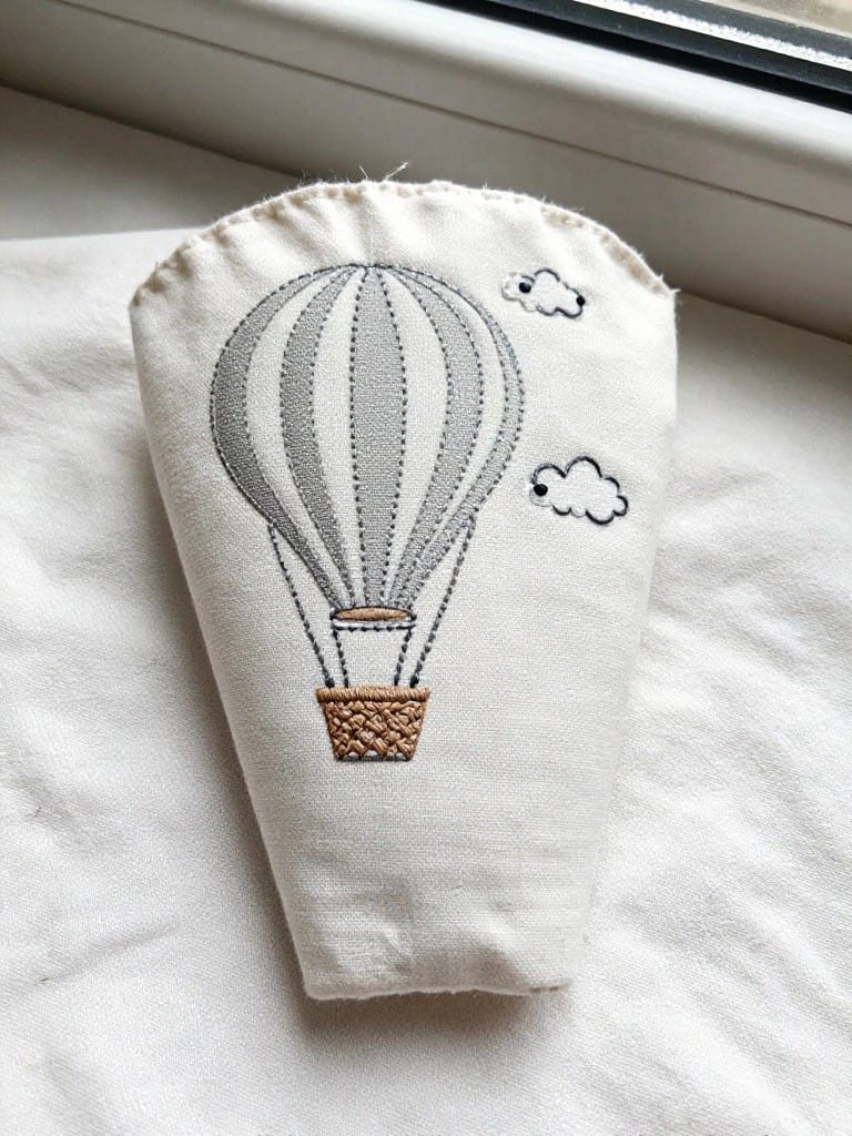White tapered fabric sleeve embroidered with gray hot air balloon, two white clouds, and brown basket.
