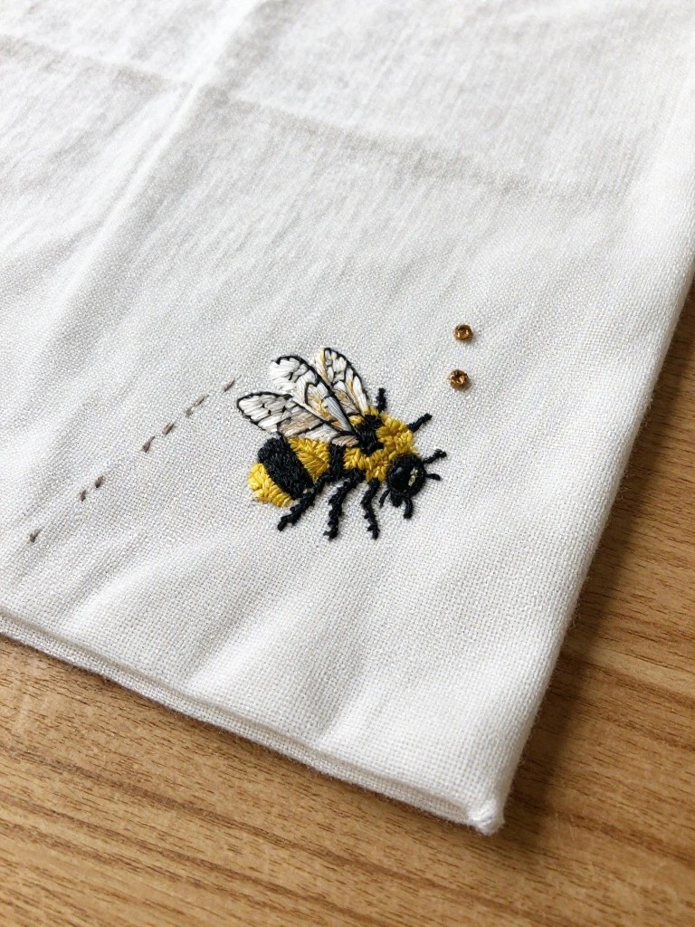 White fabric corner embroidered with a yellow and black bumblebee, dashed line, and three trailing dots on a wooden surface.