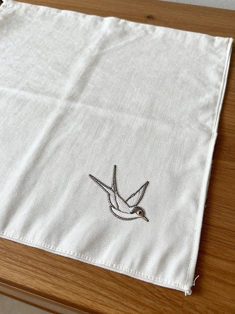 White square cloth with a small embroidered swallow bird in dark thread near the bottom edge, on a wooden table.