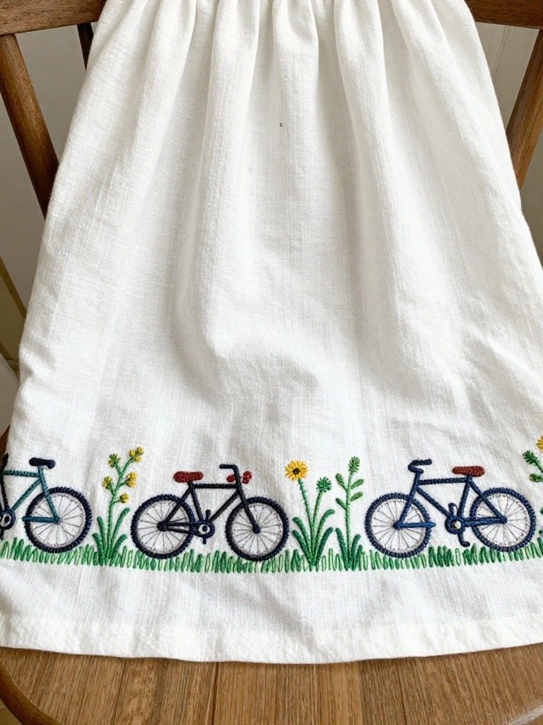 White apron draped over a wooden chair with a row of three embroidered bicycles on green grass and yellow flowers along the bottom hem.