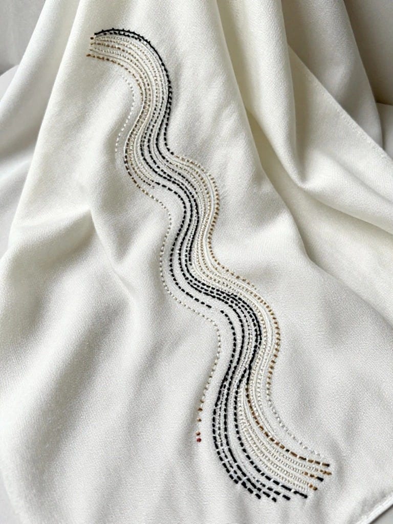 White rectangular fabric with embroidery of multiple parallel wavy lines in black, gray, and beige along one edge.