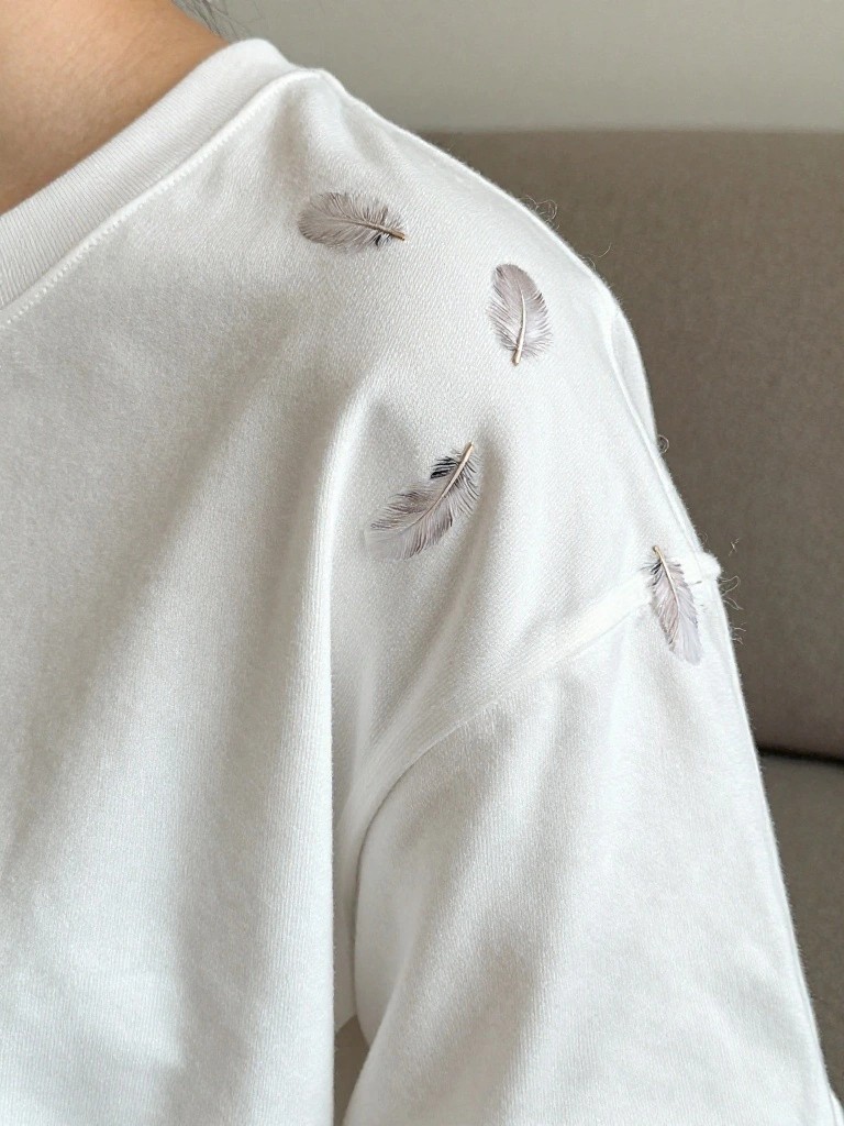 White sweatshirt shoulder with small embroidered feathers in matching light tones scattered near the seam.