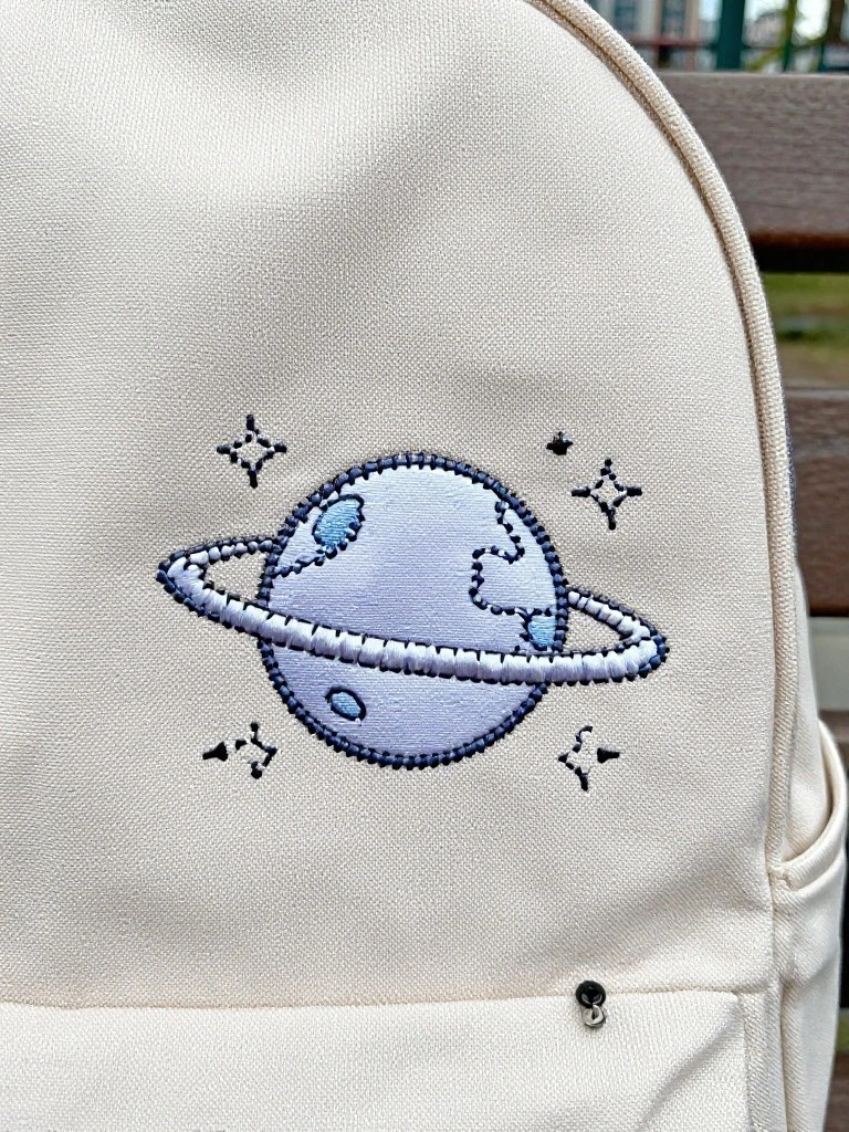 Beige canvas backpack with blue embroidered Saturn planet, rings, and stars on the back flap.