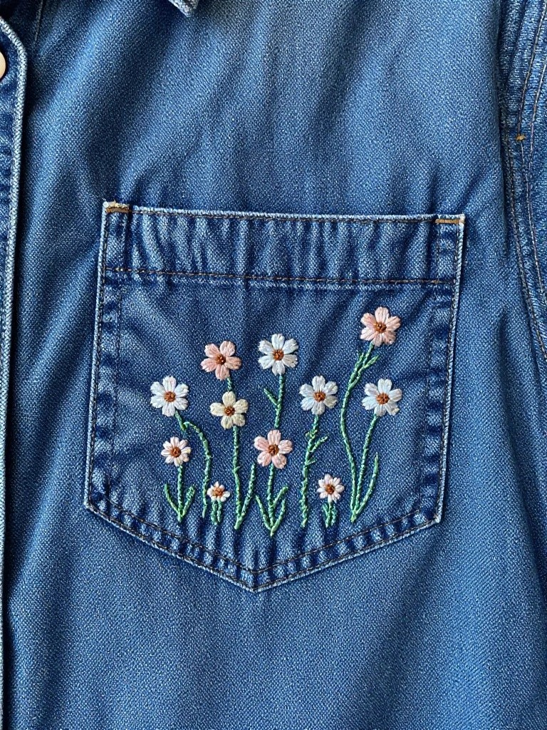 Close-up of small white and pink embroidered daisies with green stems on the pocket of a blue denim shirt.