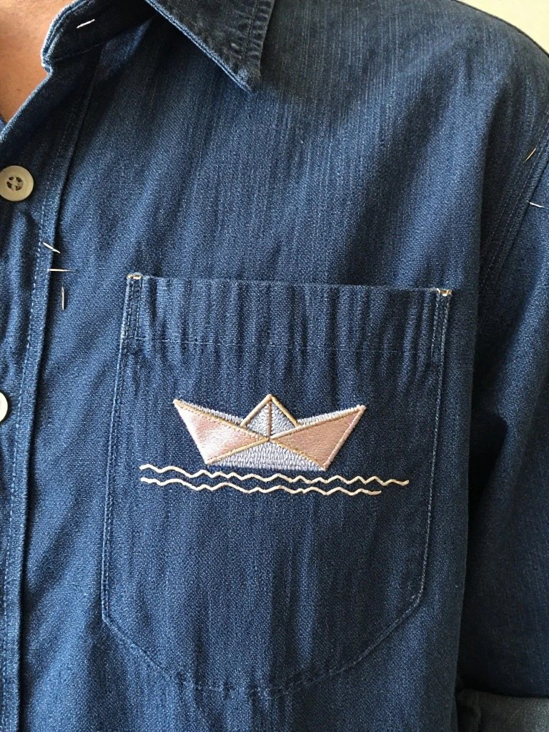 Denim shirt pocket embroidered with a pink and white origami boat motif and wavy lines below it.