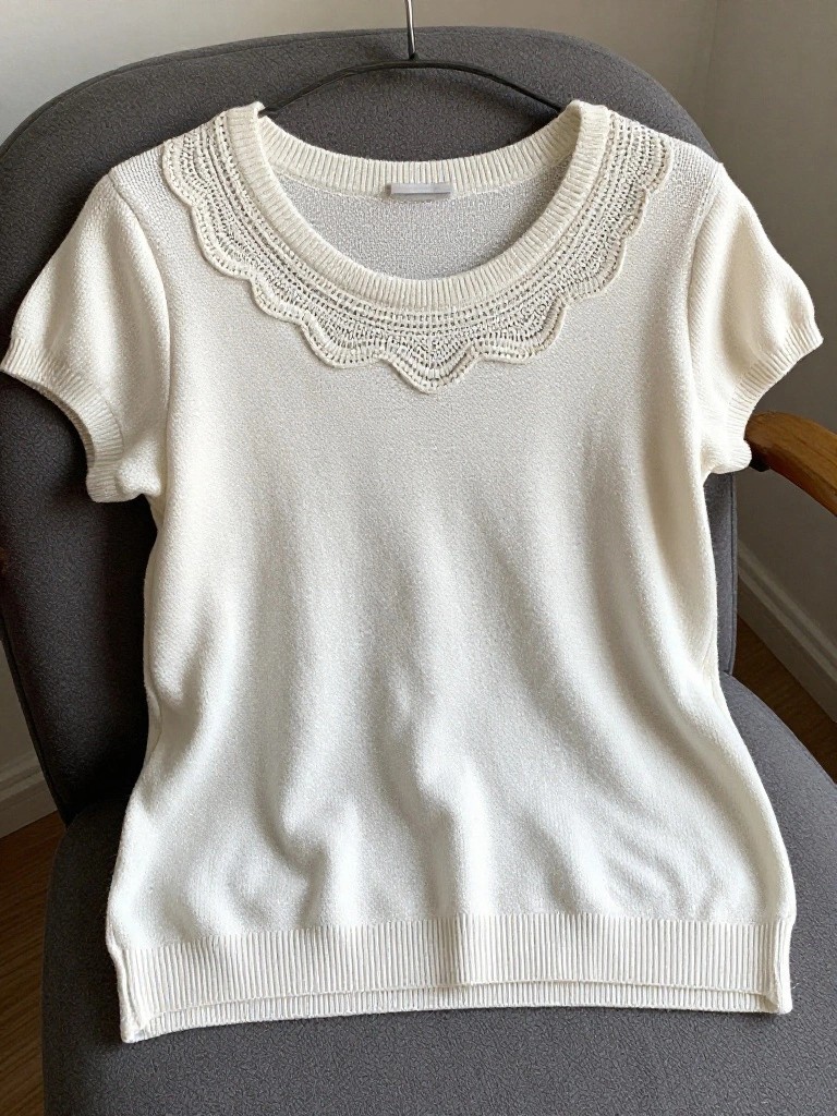 White short-sleeved knit shirt with scalloped embroidery around the crew neckline, hanging on a hanger over a gray chair.
