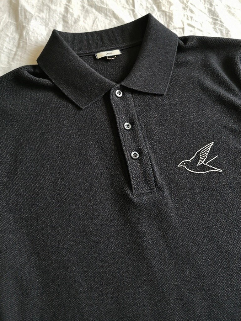 Black polo shirt laid flat with white embroidered outline of a flying swallow bird on the left chest area.