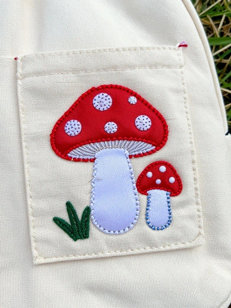 Close-up of two embroidered red mushrooms with white spots on green grass appliqued to a white apron pocket.