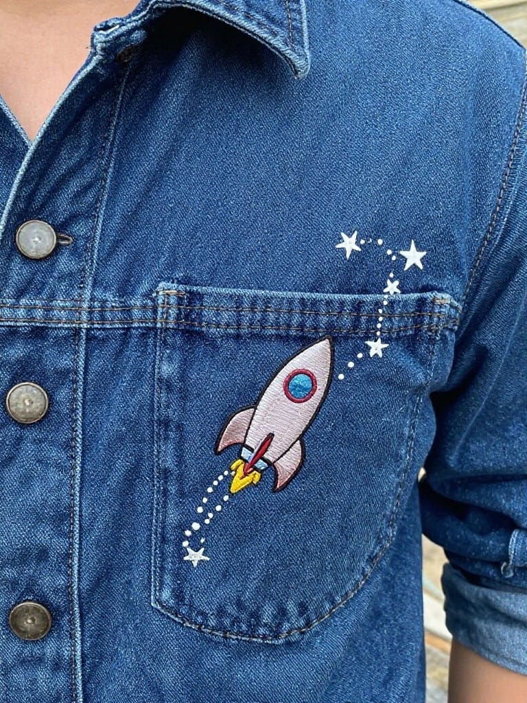 Light wash denim jacket pocket embroidered with a white rocket ship emitting yellow exhaust and trailed by white stars.