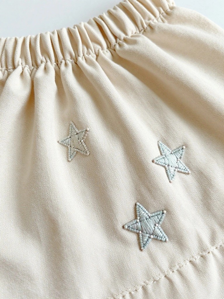 Close-up of beige gathered shorts with several small light blue embroidered stars scattered across the front lower fabric.
