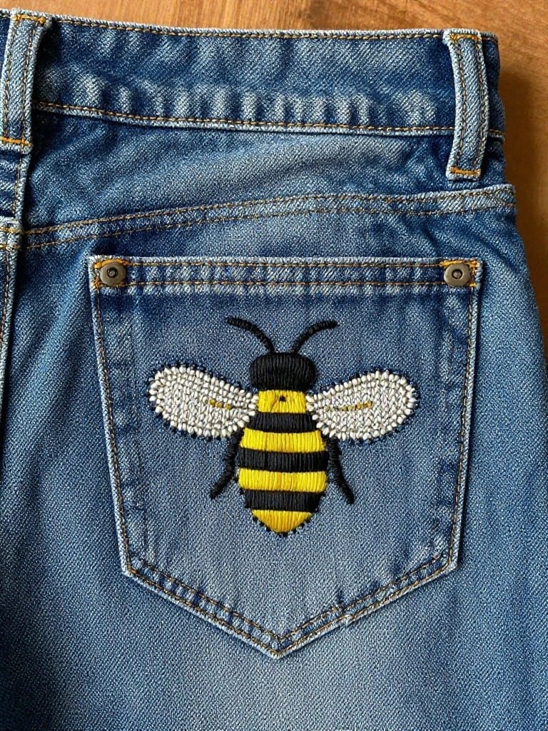 Yellow and black embroidered bee with sparkly wings on the back pocket of blue jeans.