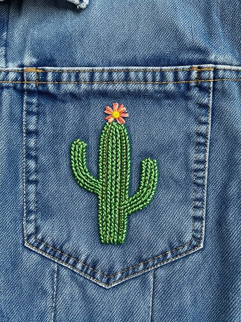 Green embroidered cactus with pink flower on the pocket of a blue denim jacket.