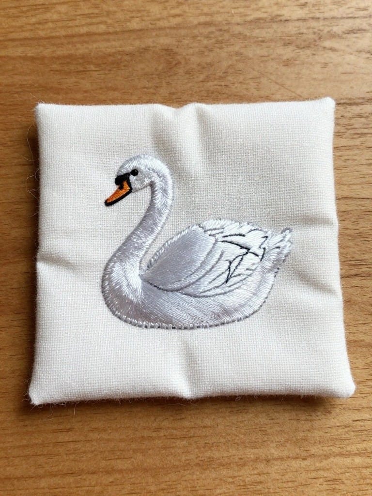 Small white stuffed fabric square embroidered with a white swan that has an orange beak.