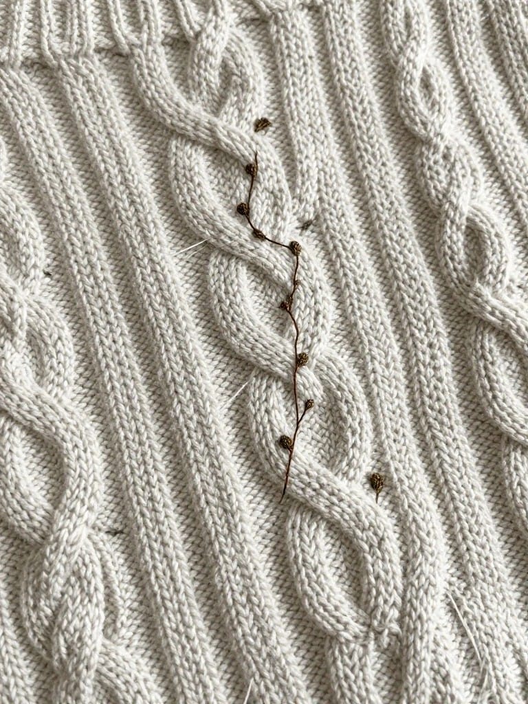 Close-up of cream cable knit sweater fabric with a small embroidered brown berry stem following one cable twist.