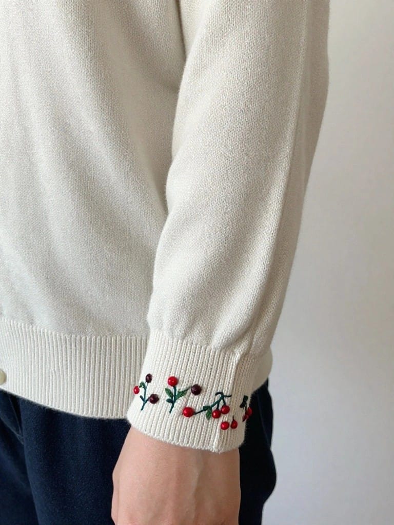 Cream sweater sleeve with red cherry and green leaf embroidery along the cuff, held by a hand.