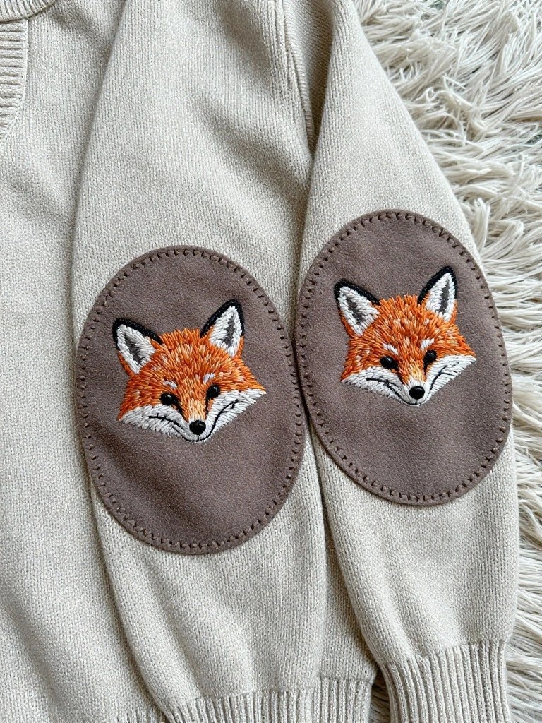 A cream knit sweater with oval elbow patches featuring embroidered fox faces in orange and brown tones.