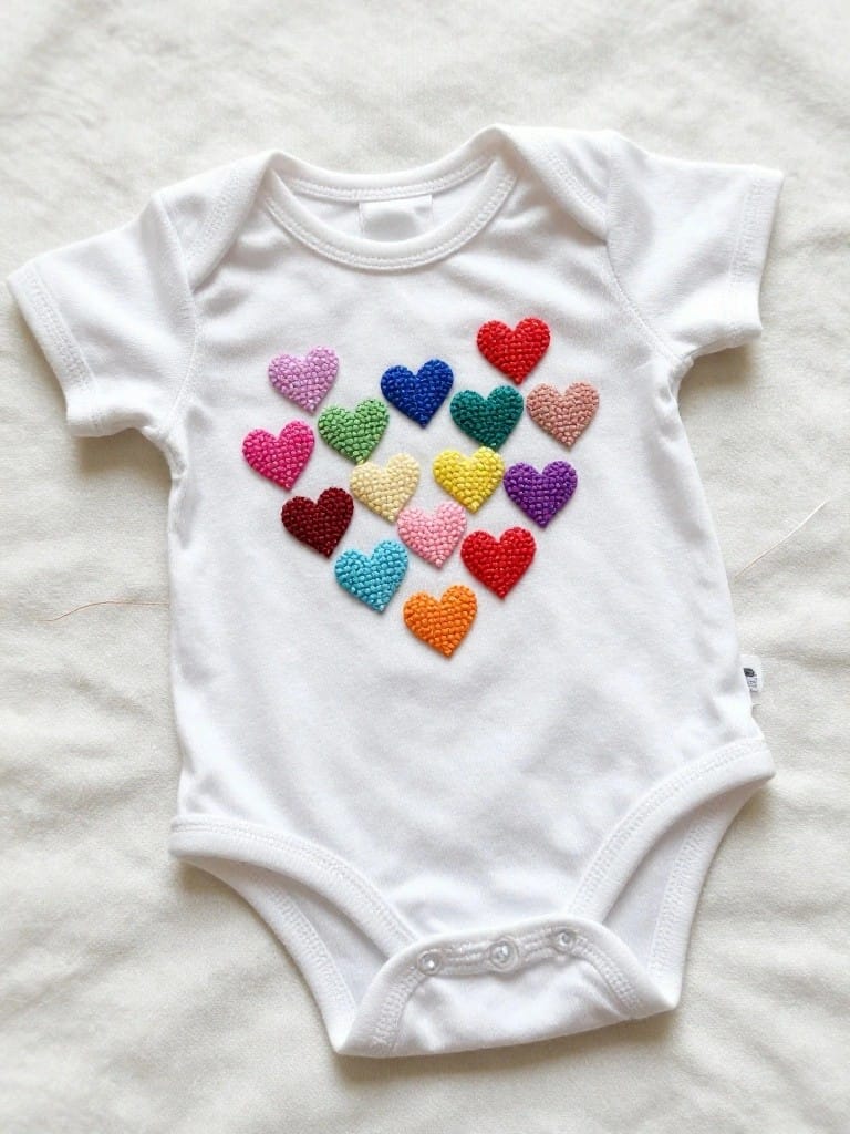 White short-sleeve baby onesie with about a dozen small colorful fabric hearts sewn into a larger heart shape on the chest.