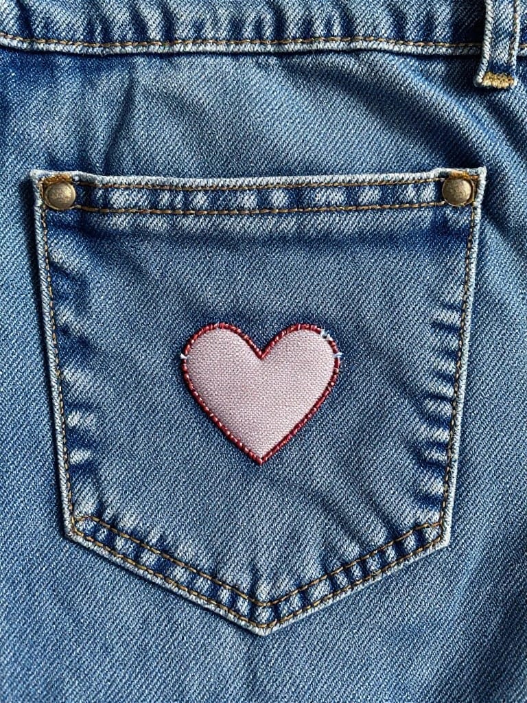 Blue denim jeans pocket flap with a pink fabric heart appliqued on top and stitched around the edges.