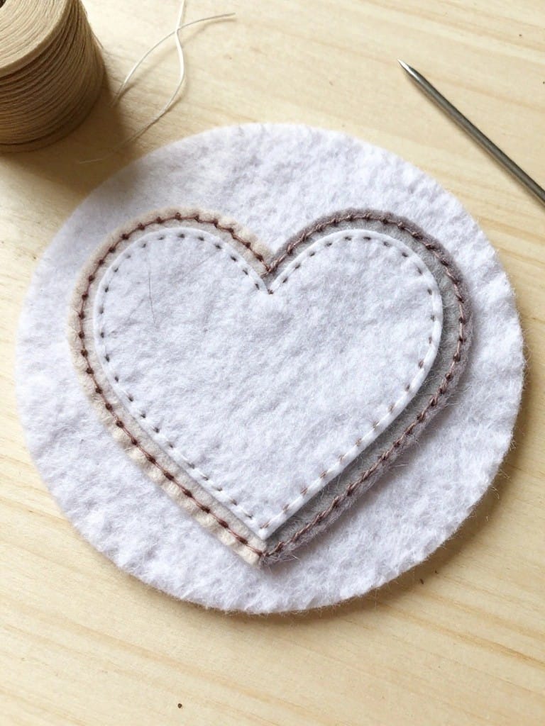 White felt circle with double brown-stitched heart outline on wooden surface, brown thread spool and needle nearby.