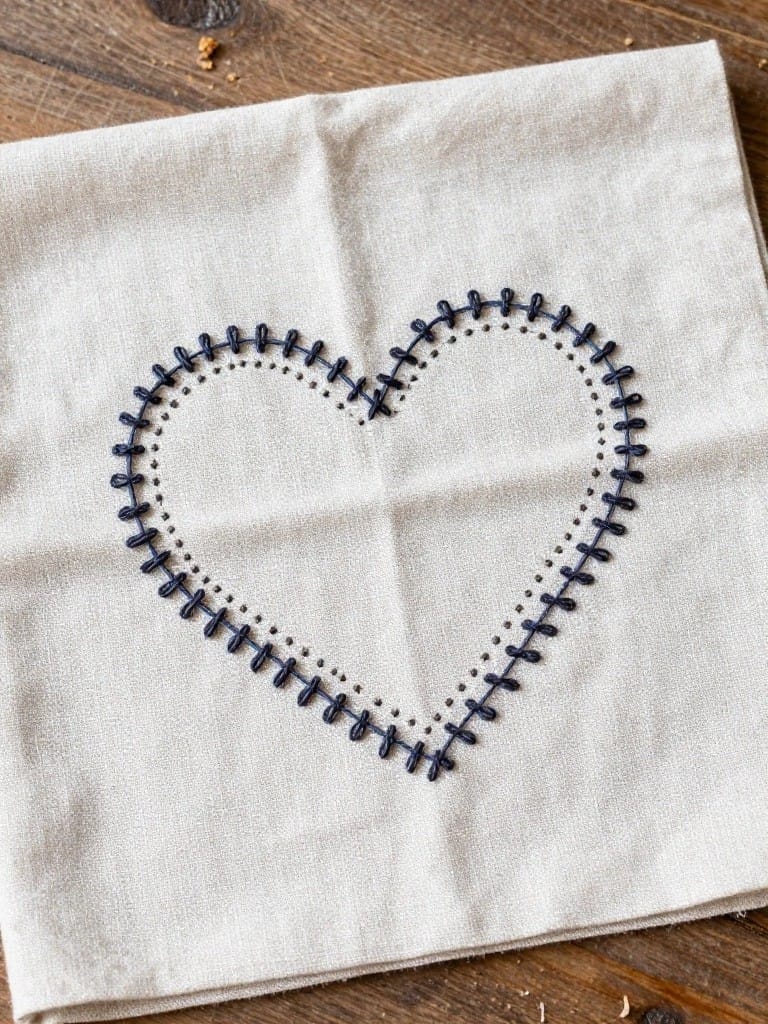 White square linen napkin with a large navy thread outline heart stitched in the center, folded on a wooden table.