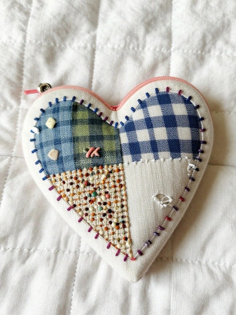 A heart-shaped fabric pouch made from patchwork squares in blue plaid, green check, and other patterns, with embroidered edges, beads, and a pink zipper.