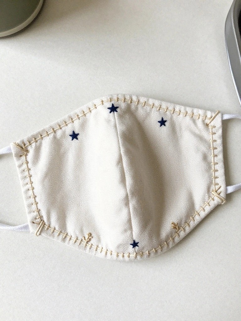 White cloth face mask with navy embroidered stars near the edges and stitched borders in off-white thread.