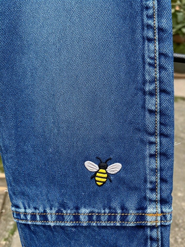 Close-up of yellow and black embroidered bee on lower leg of blue jeans near hem.