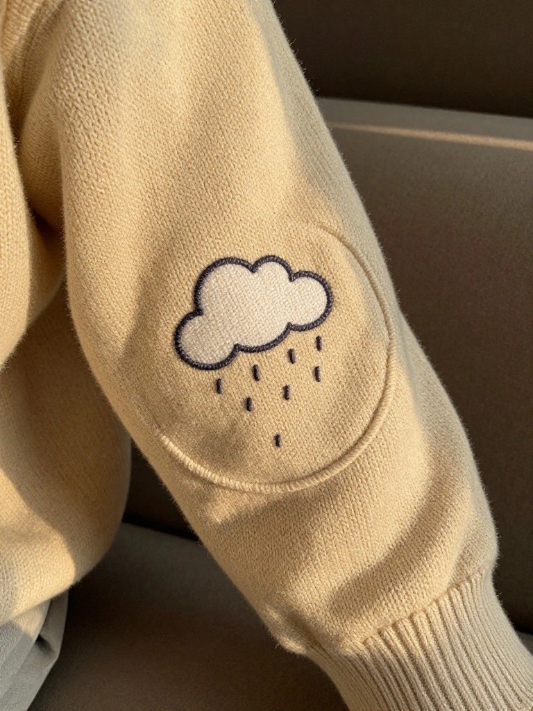 Cream knit sweater sleeve with dark thread embroidered cloud and raindrops in a round elbow patch.