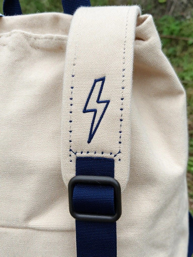Beige canvas tote bag with navy strap featuring an embroidered navy lightning bolt near the black buckle.