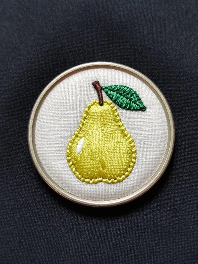 A small metal embroidery hoop displays a yellow embroidered pear with green leaf on white fabric against a black background.