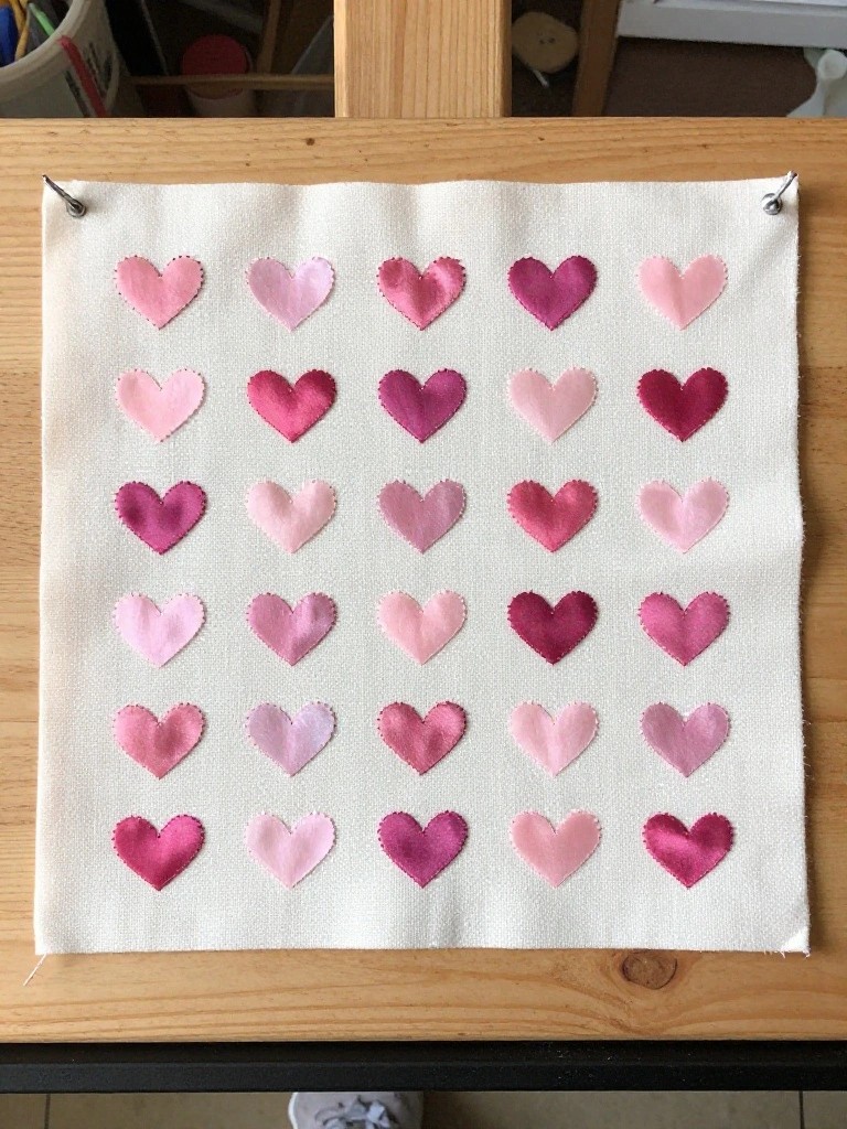 White fabric square with a 6 by 6 grid of small appliquéd hearts in shades of pink and red, stretched on a wooden frame.