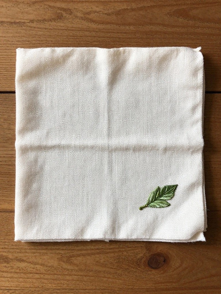 White napkin folded into quarters on a wooden table with a small green embroidered leaf in one corner.