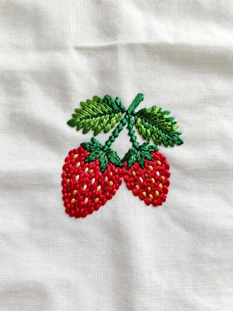 Two red embroidered strawberries with green leaves on white fabric.