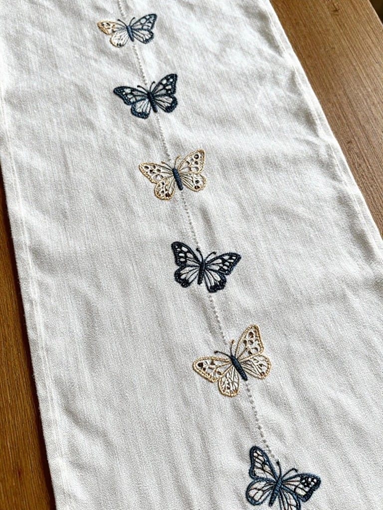 White linen table runner with navy and gold embroidered butterflies spaced down the center on a wooden table.