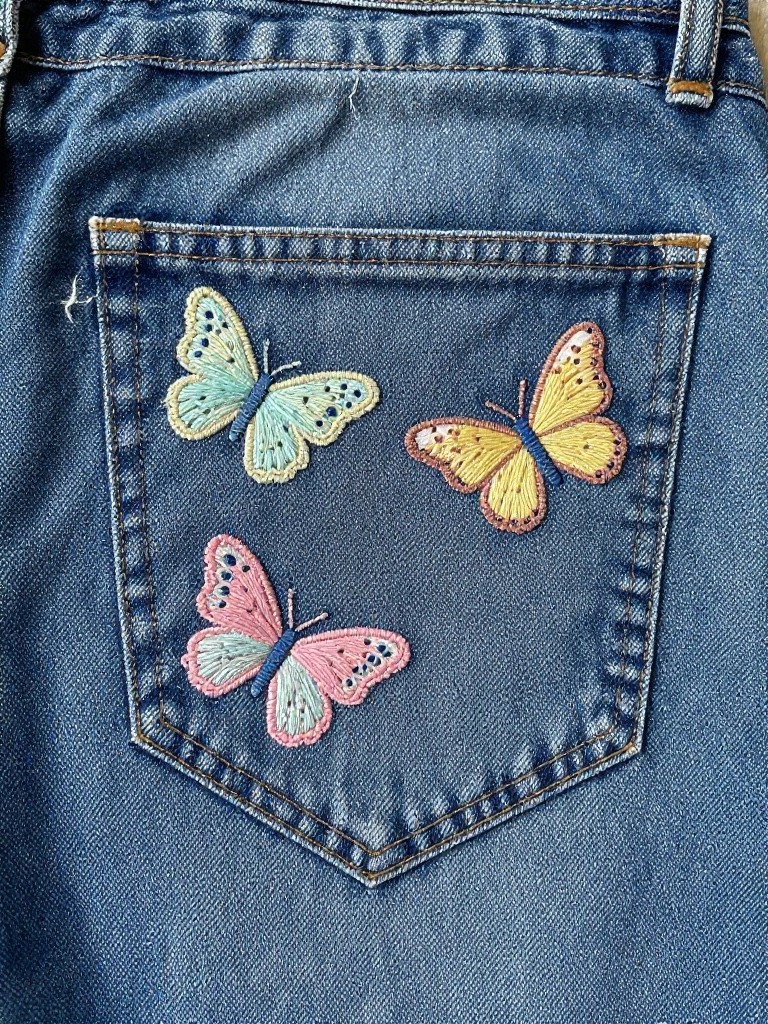 Back pocket of blue jeans with three embroidered butterflies in light blue, yellow-orange, and pink.
