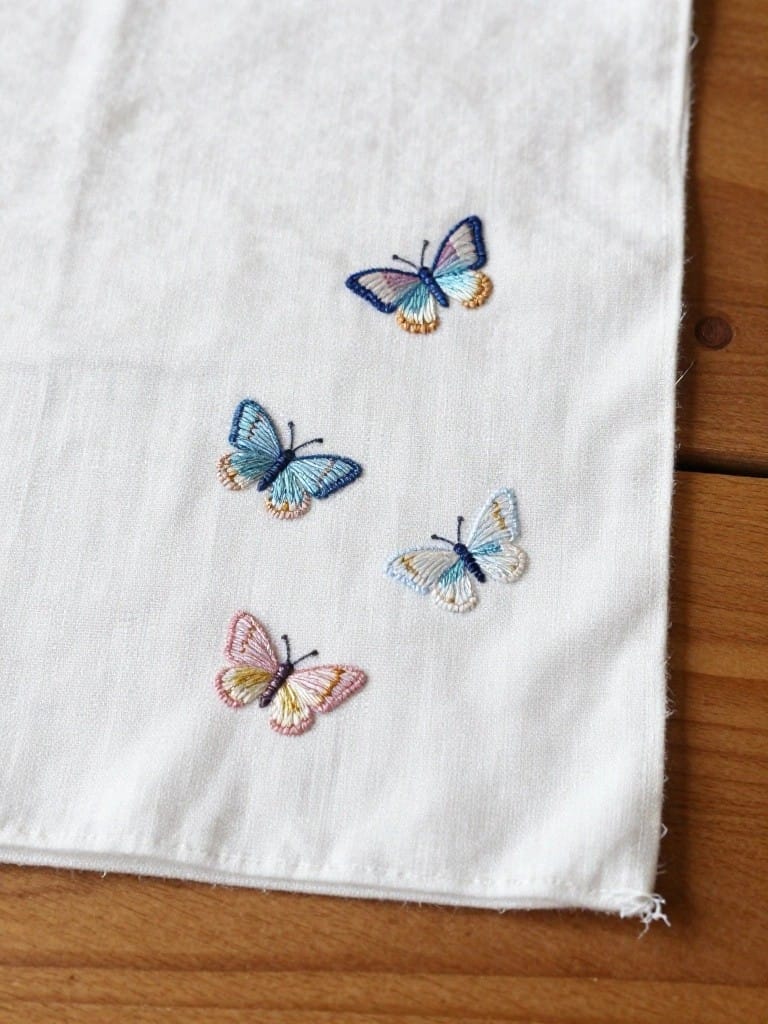 White napkin embroidered with four small butterflies in blue and pink shades scattered near the bottom edge on a wooden table.