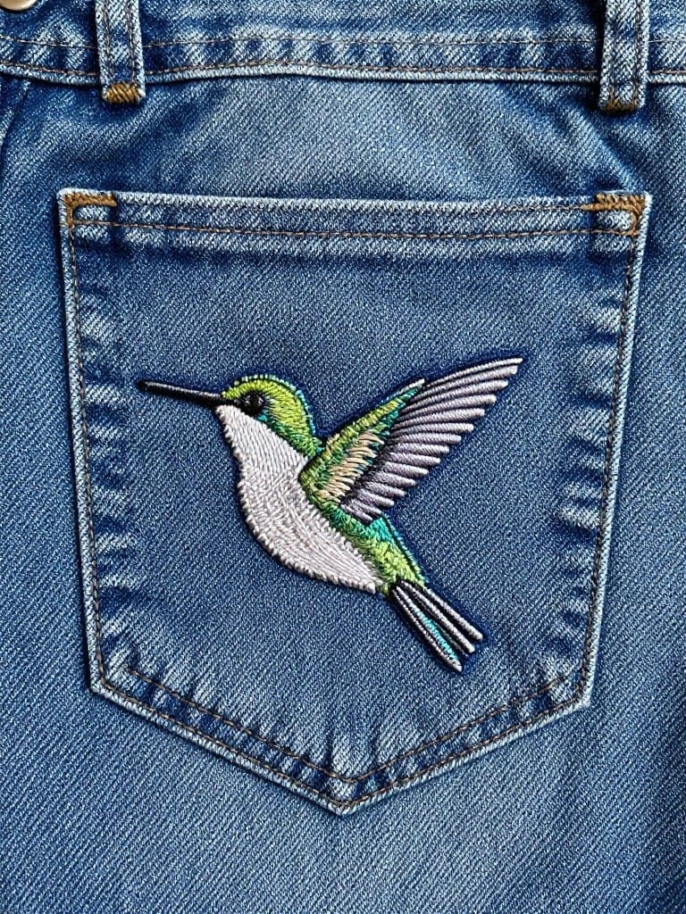 Back pocket of light wash jeans embroidered with a green, white, and blue hummingbird.