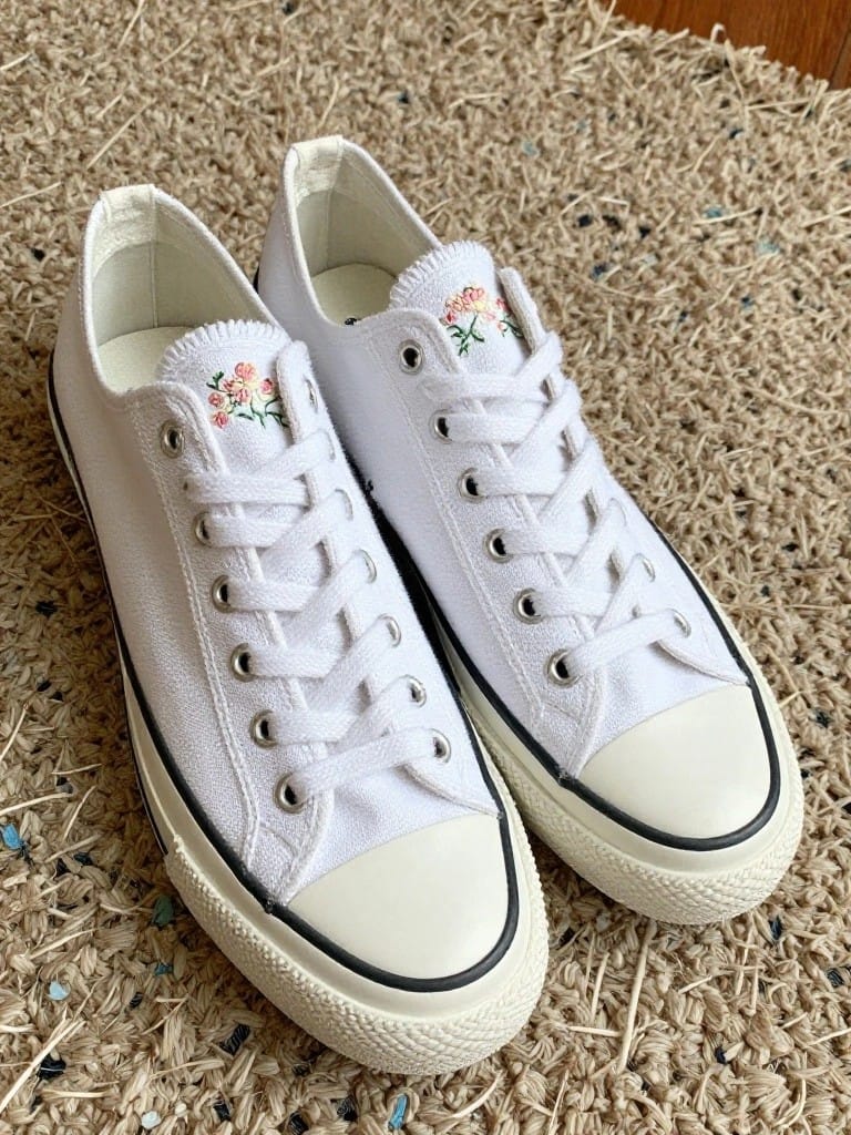 Pair of white canvas sneakers with pink embroidered flowers and green leaves on the outer side panels.