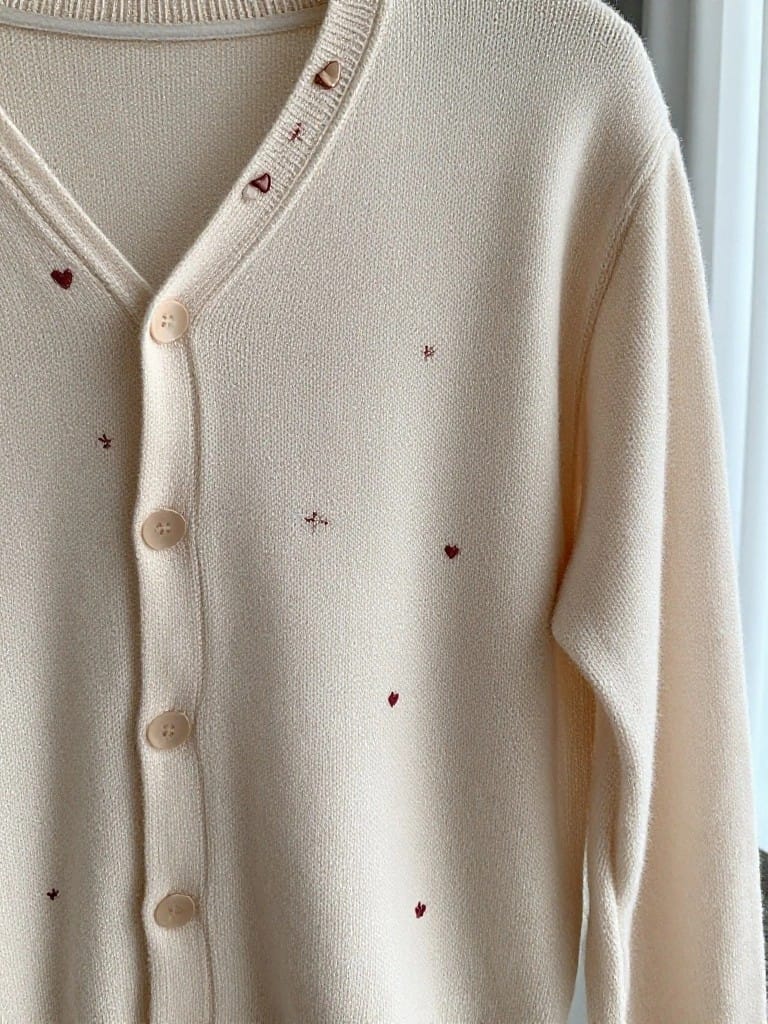Cream v-neck cardigan with small red embroidered hearts, stars, and crosses scattered across the front panels and collar.