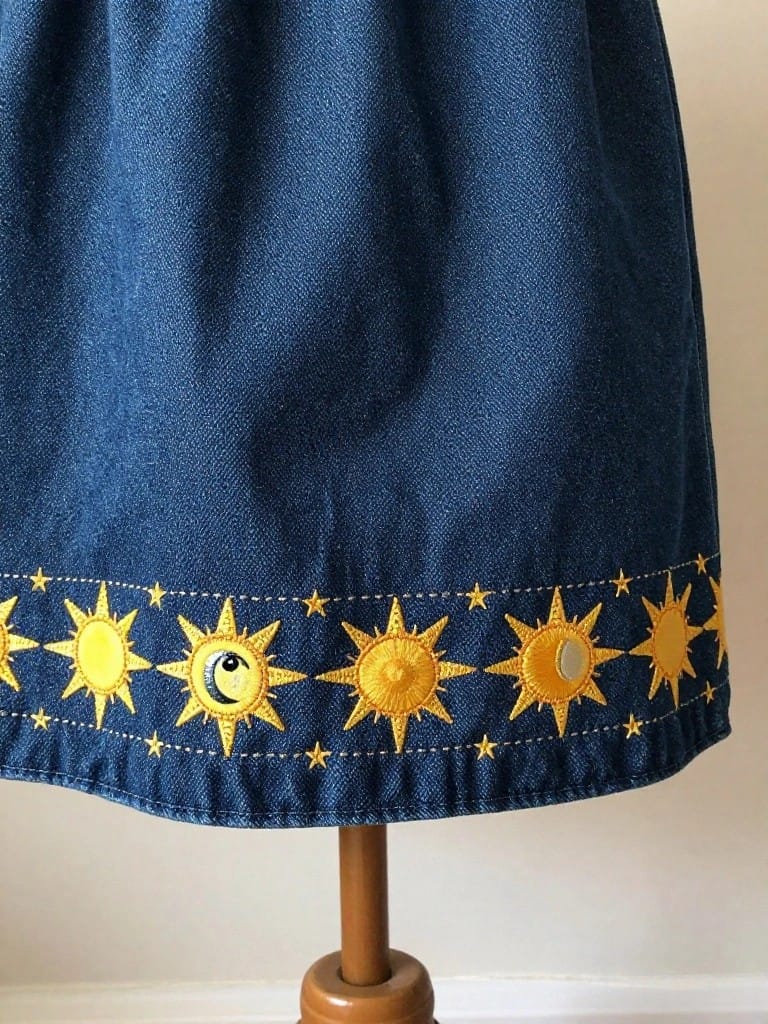 Navy blue denim skirt with yellow embroidered suns, crescent moons, and starry elements along the bottom hem, shown on a wooden stand.