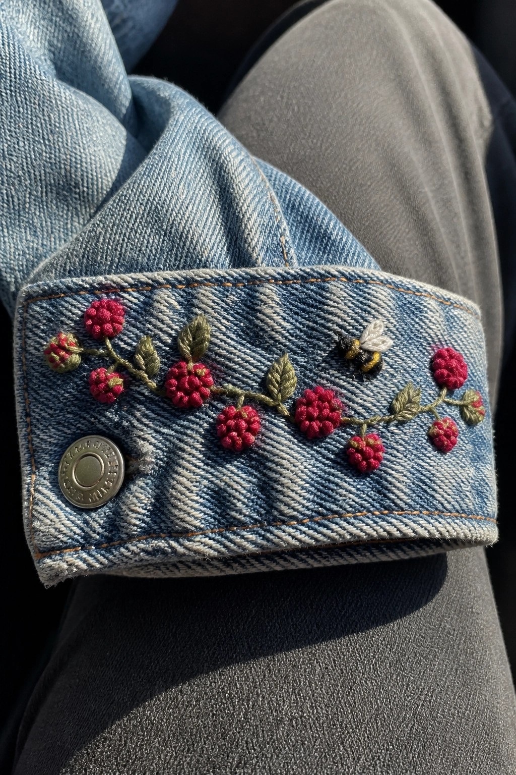 Blue denim jacket cuff embroidered with red berries, green leaves, and bee.