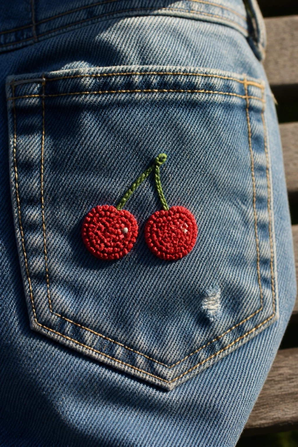 Red French knot cherries with green stems embroidered on a blue denim jeans pocket.