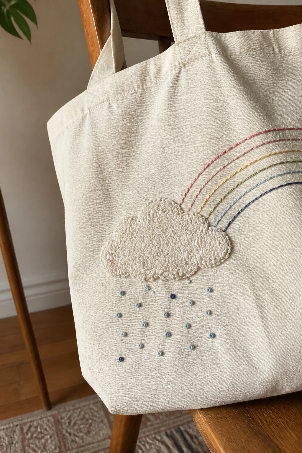 Embroidered cream tote bag with cloud, rainbow, and blue raindrops on wooden chair.