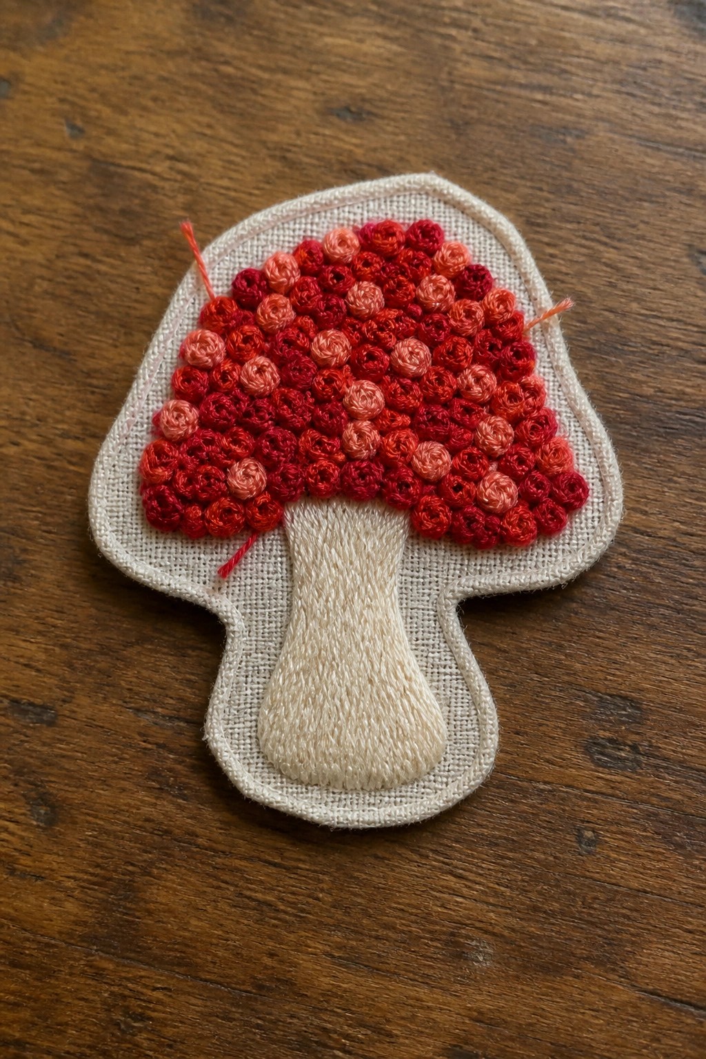 Mushroom-shaped fabric patch with red and orange French knots filling the cap and plain beige stitching on the stem.