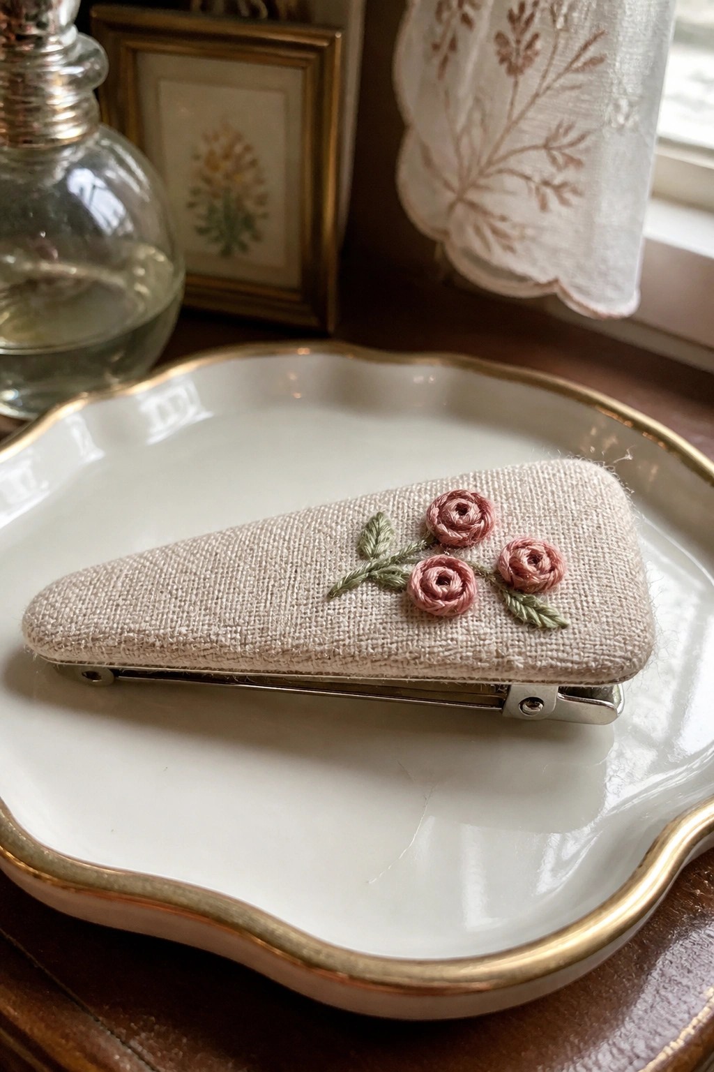 Beige fabric hair clip with pink rose embroidery on white gold-rimmed dish.