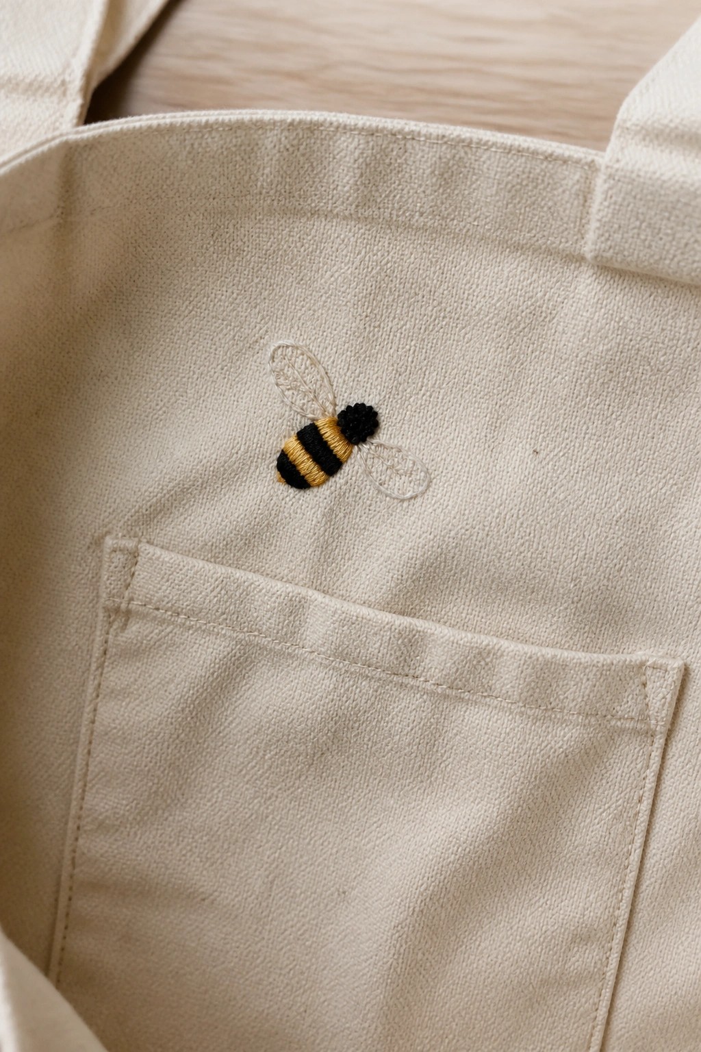 Beige canvas tote bag with embroidered yellow and black bumblebee above pocket.