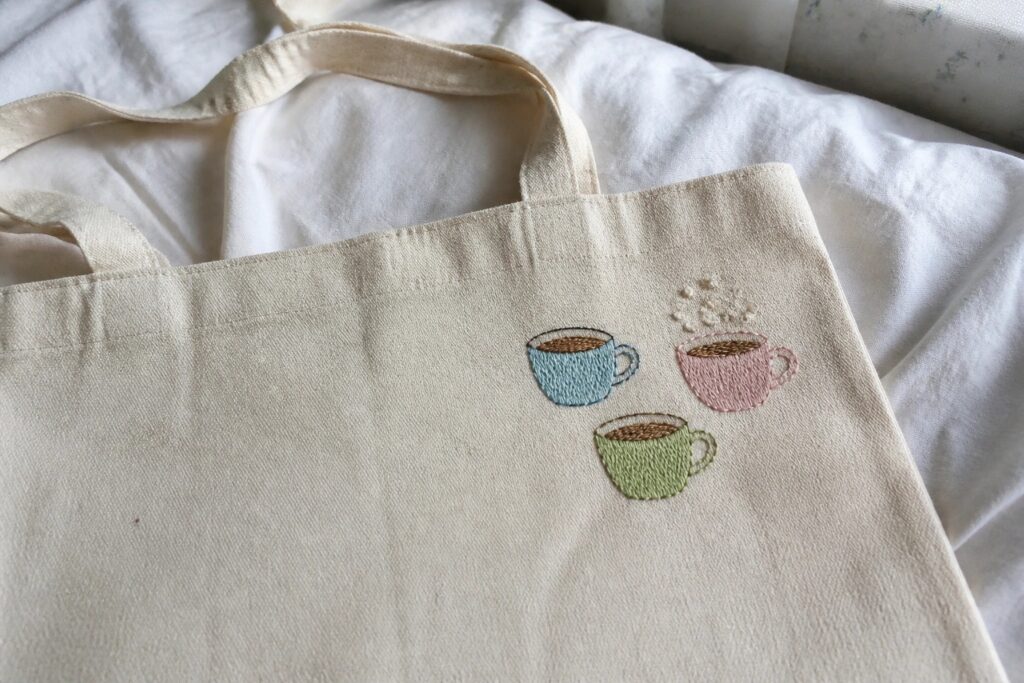 18 Cute Tote Bag Embroidery Ideas for Everyday Errands, Books, and Coffee Runs Three embroidered coffee cups in pink, green, and blue on a cream-colored tote bag.