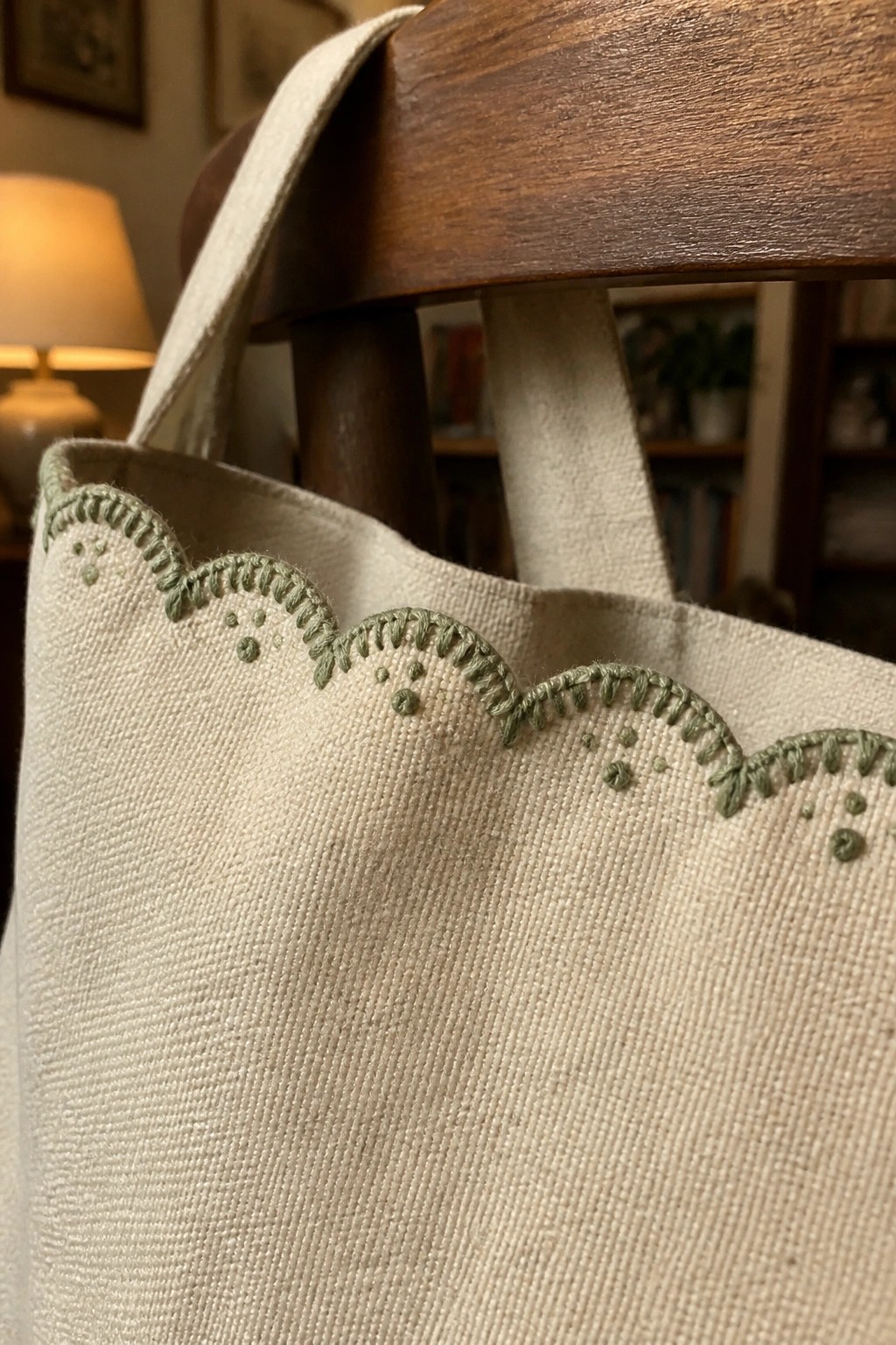 A close-up of a beige canvas tote bag with green scalloped embroidery along the top edge resting against a wooden chair.