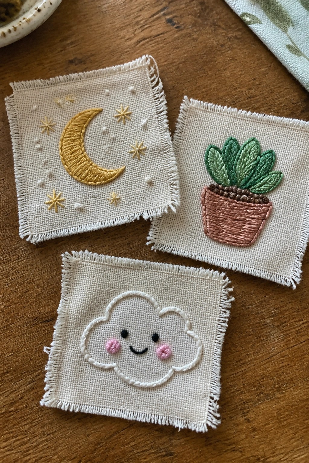 Three small fabric squares with embroidered moon and stars, potted plant, and smiling cloud designs on a wooden surface.