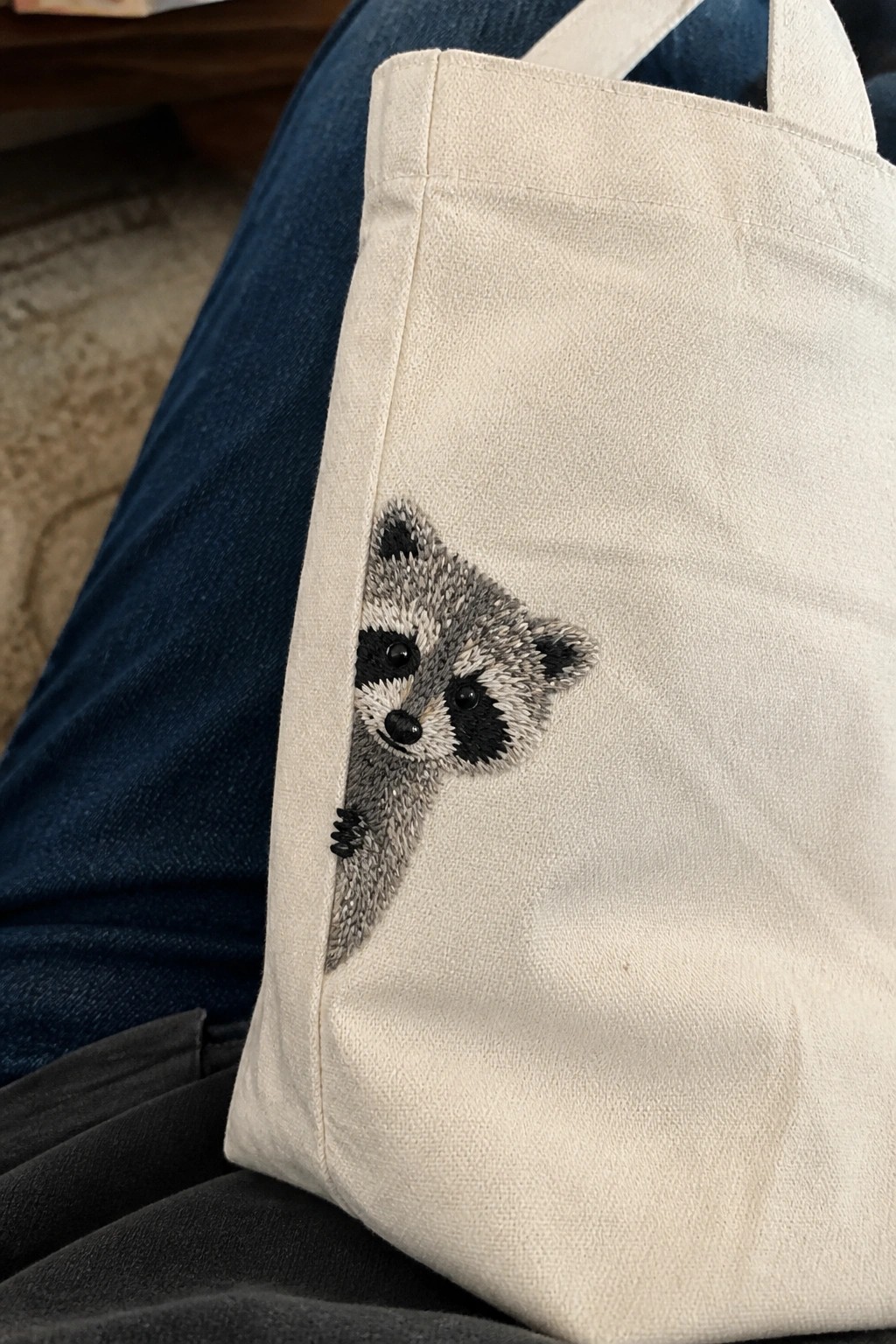 A raccoon head and paw embroidered on the side of a white canvas tote bag, positioned to look like the animal is peeking from the seam.